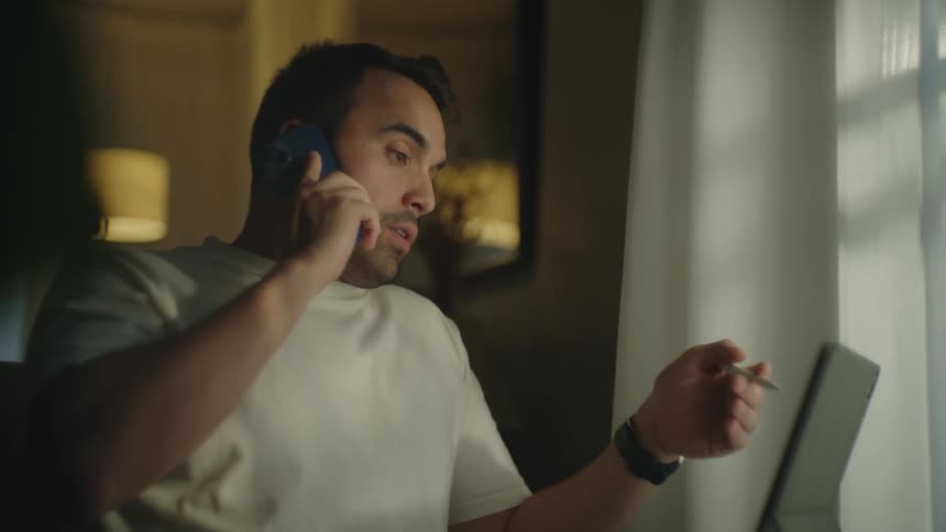 Man sitting in room consults laptop screen while talking on phone