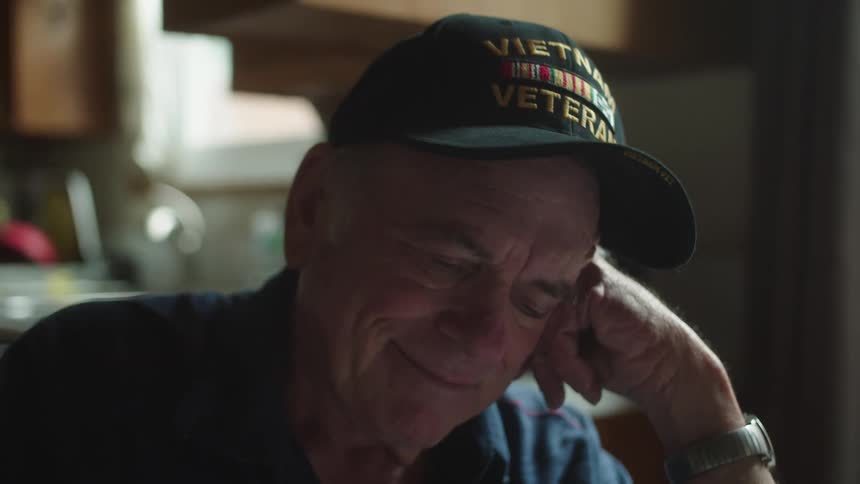 Elderly man sitting near kitchen and wearing vietnam veteran hat smiles while reading