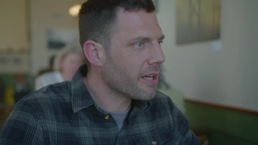 Man sits in busy restaurant talking to companion sharing his table