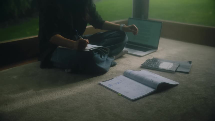 Woman sits cross legged on floor near windows with computer and works on schoolwork