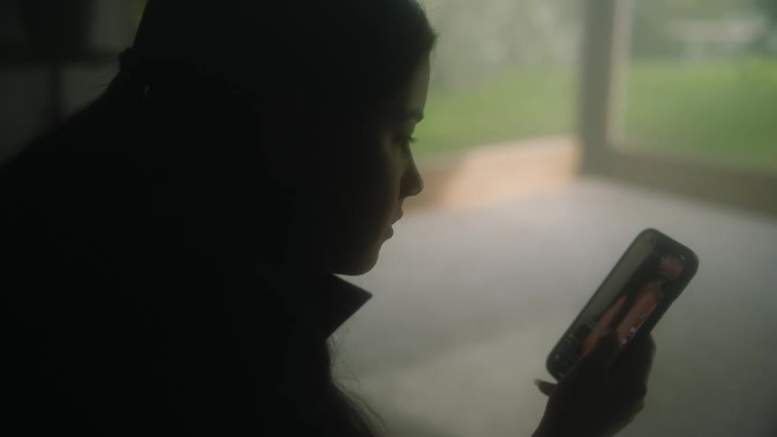 Woman swiping down her smartphone is silhouetted against the windows overlooking backyard