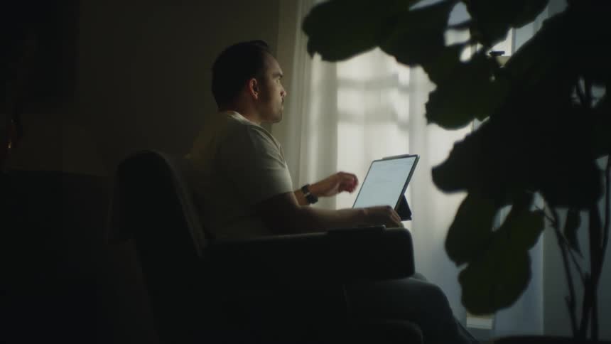 Man sitting in dim room types on computer keyboard then pauses to think