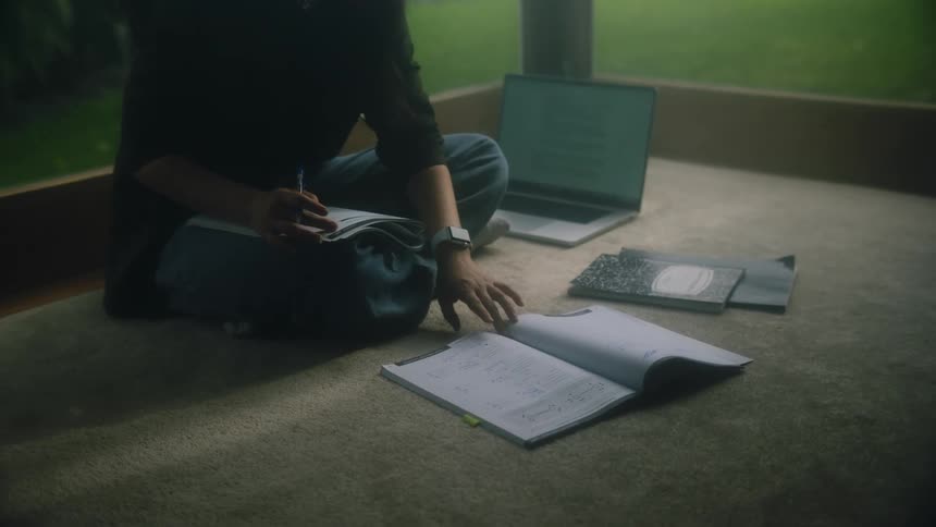 Woman sits on floor near windows working on homework