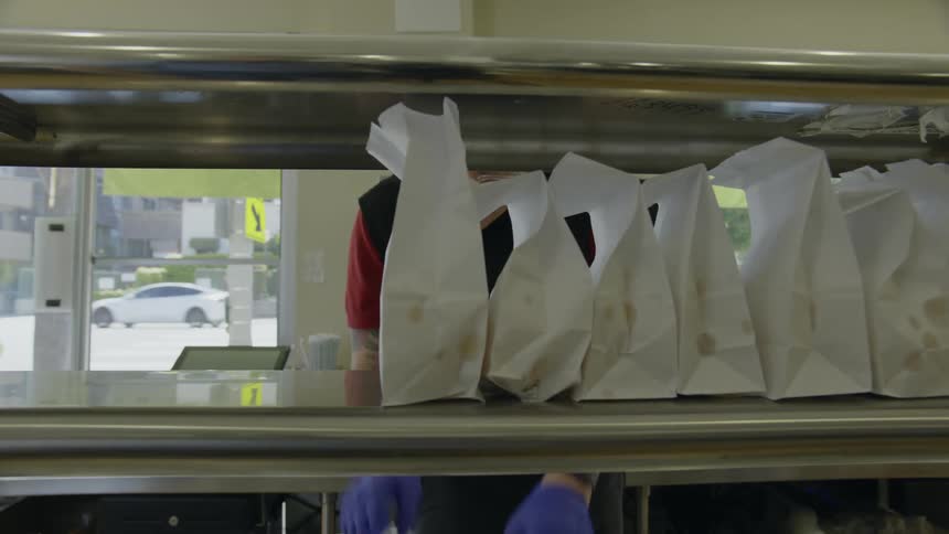 Restaurant worker places filled food bags onto metal shelves as traffic passes on road in the background