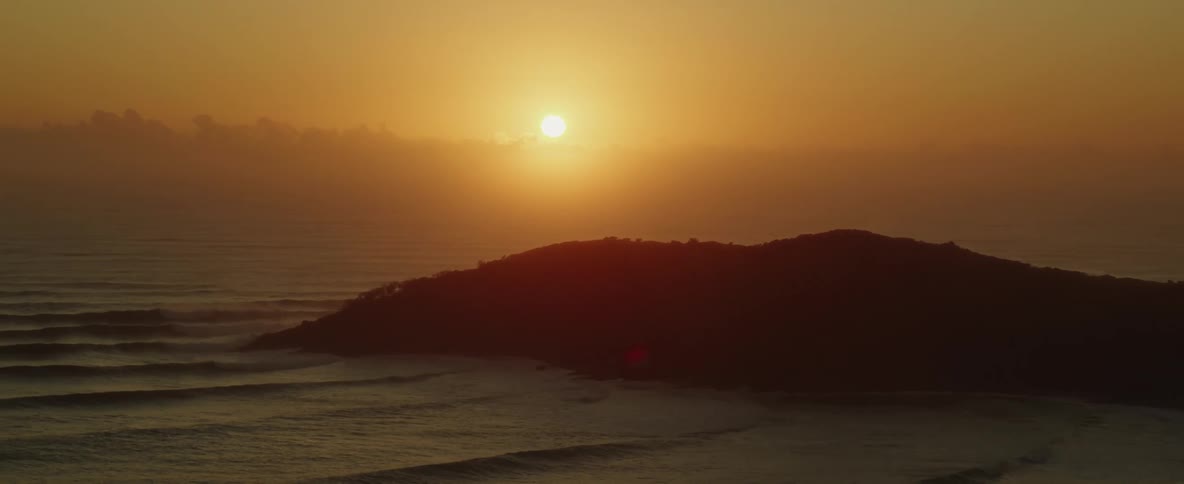An aerial of the sun setting in the evening above the silhouette of a coastal hill as waves roll onto shore