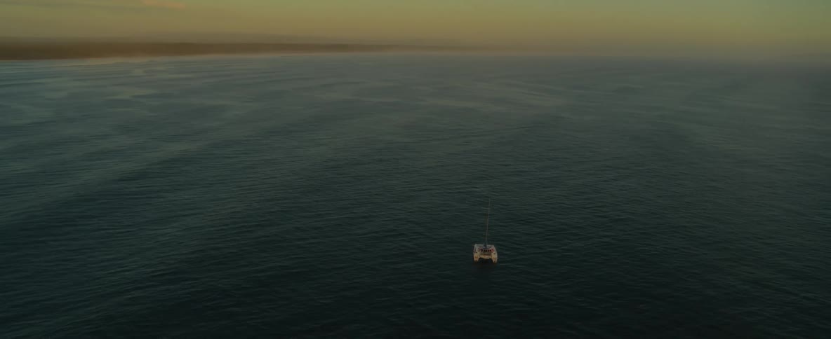 Catamaran boat floats on ocean a distance from the shore under an amber sky