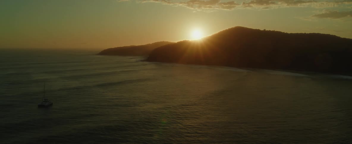 An aerial of the ocean waves crashing against the silhouette of a hillside as the sun rises over it