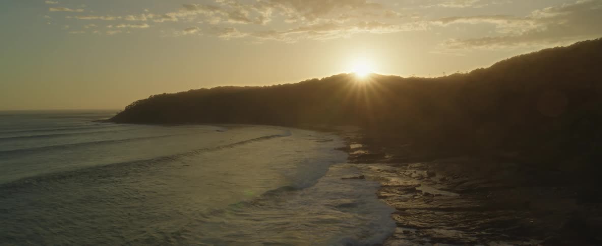 The silhouette of hills as the sun sets behind them as waves roll onto the beach