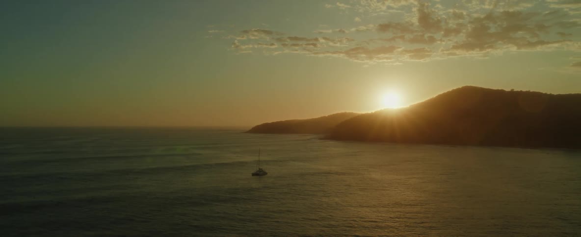 An aerial of a boat on the ocean floating on the water near a mountain on the coast as the sun sets behind it