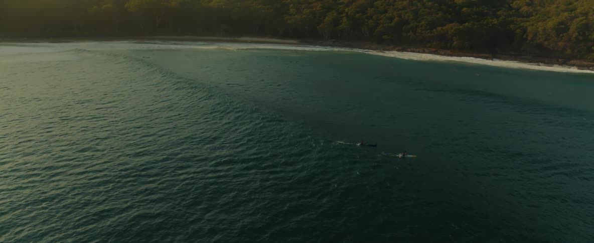 Sunlight sparkles on the water as people in canoes ride the wave into shore and a promontory covered with trees