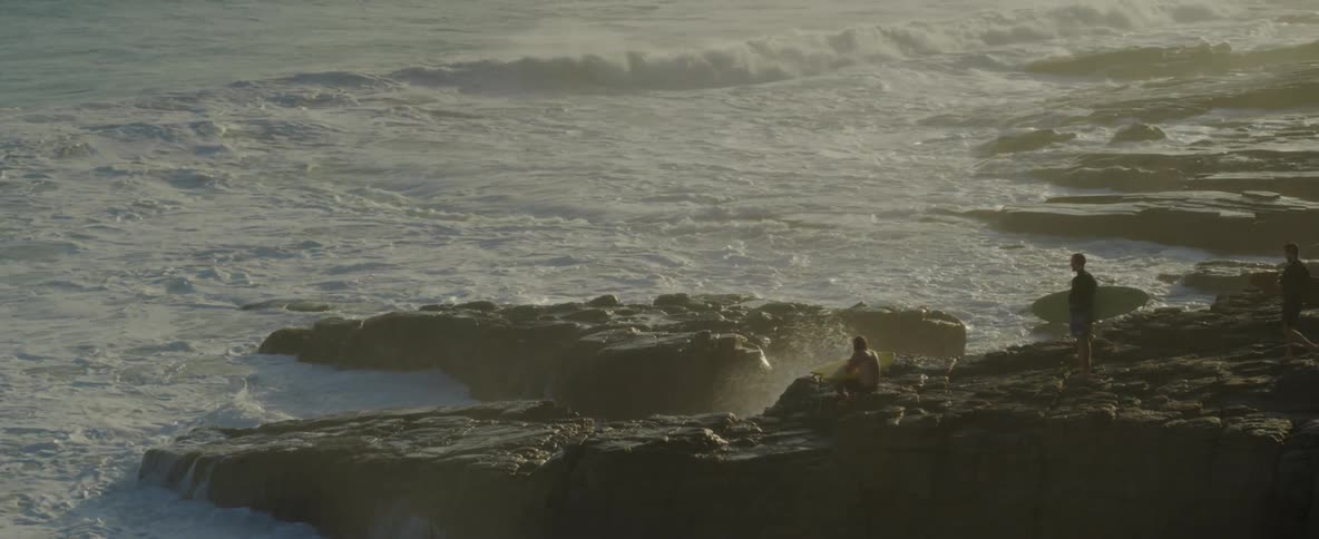 Three people with surfboards are standing on the rocky coast watching the waves roll in to shore as the man on the edge of the rock jumps in and swims on his surfboard
