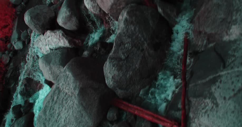 A stream of water flows down huge boulders and rocks into some red sand or grass