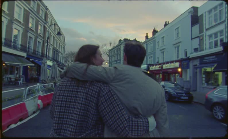 Couple walks down road with arms around each other passing storefronts toward red tinted clouds