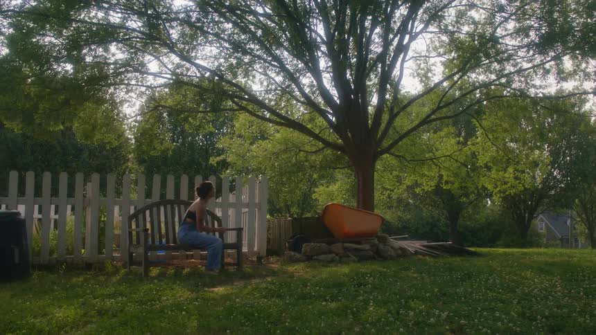 Woman walks across lawn to sit on bench with a beverage