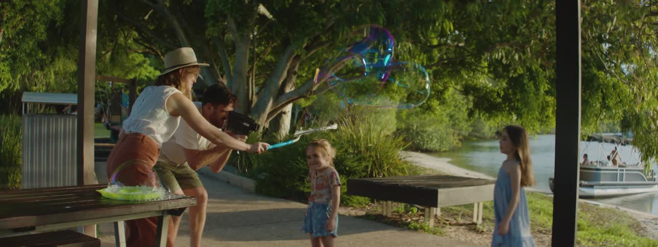 A family outdoors near a lake as the father records his daughters jumping as the mother creates bubbles