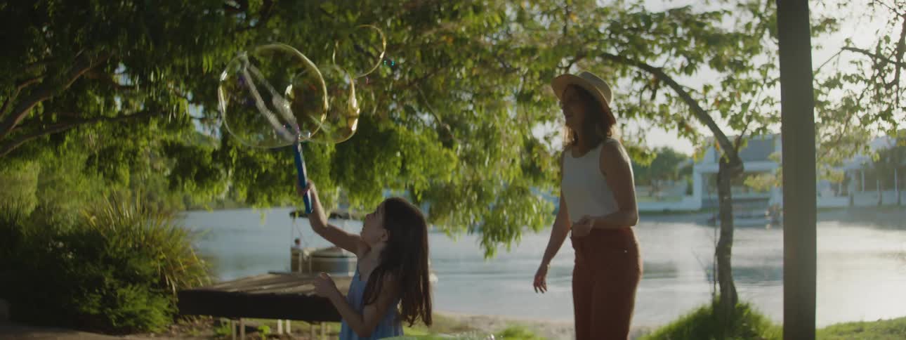 A young girl makes giant soap bubbles using a hoop while her mother watches and tries to catch them theres a lake behind them