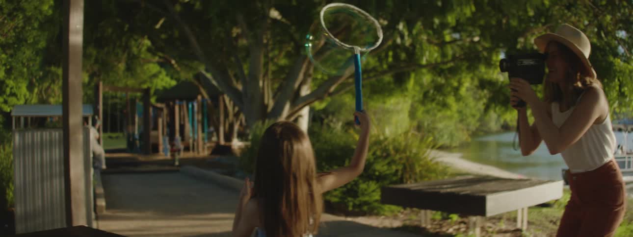 A young girl by a lake uses a hoop to make a giant soap bubble while her mother films her