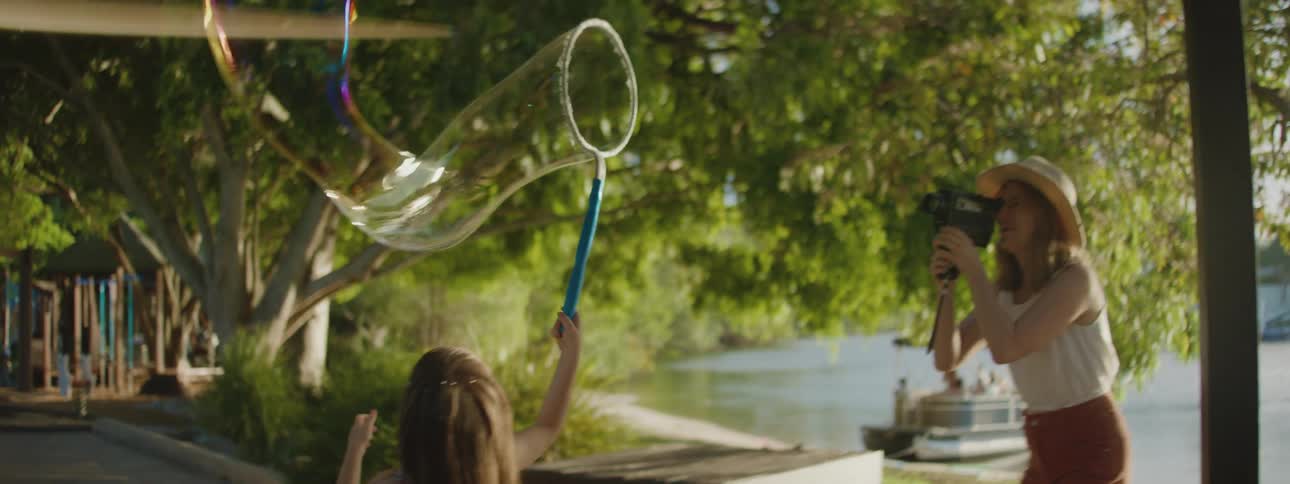 Mother with hat white top and brown pants films daughter playing with soap bubble ring near the lake