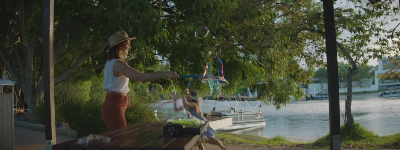 Mother and daughter play with large bubble wand as father sits on pier in the background