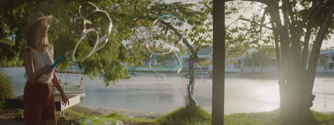A woman waving around a bubble stick creating bubbles outdoors near a lake with boats on it