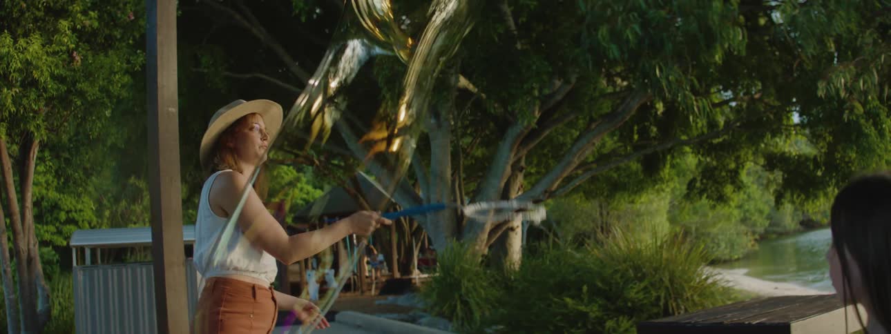 A woman next to a lake uses a big hoop to create giant soap bubbles for her young daughter