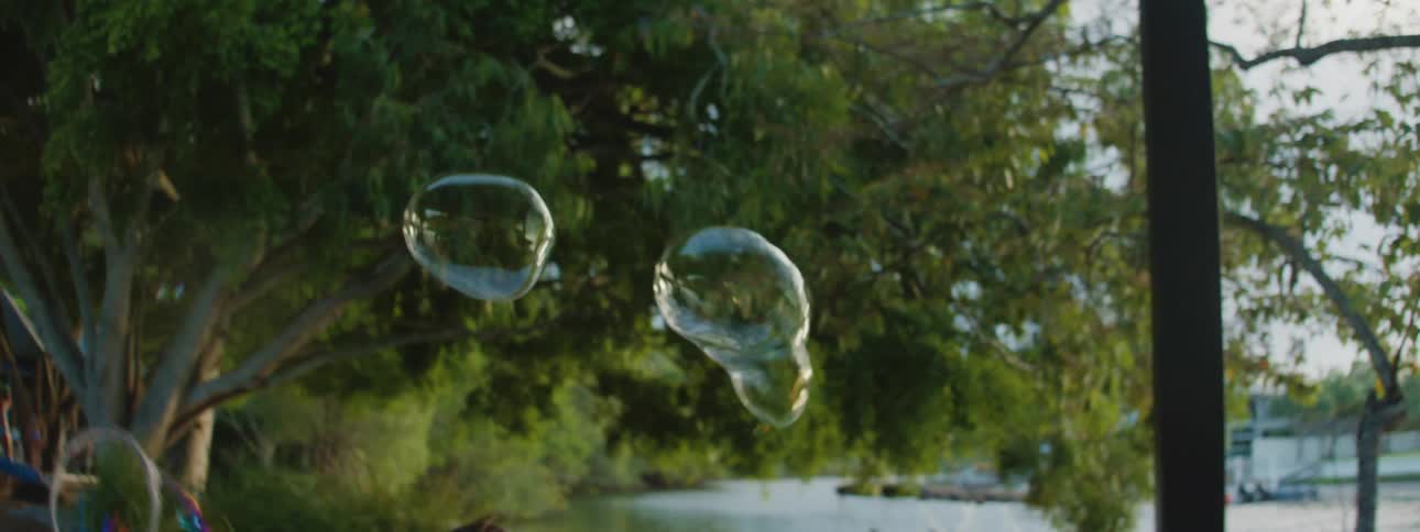 A mother by a lake uses a hoop to make soap bubbles which her young daughter then runs to try and catch