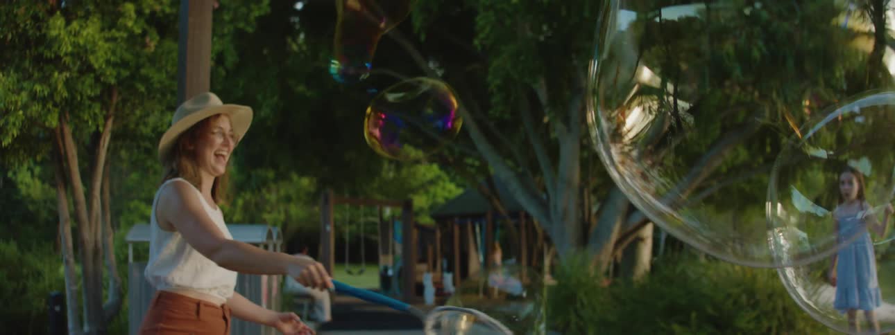 A mother makes giant soap bubbles and laughs while her daughter watches by a lake