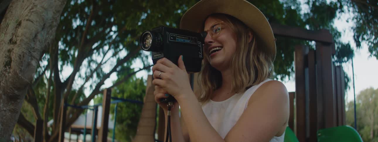 A woman wearing a sunhat is standing in a park holding a video camera as she smiles and speaks
