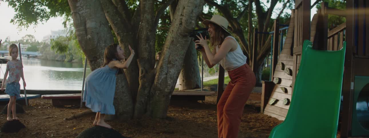 A little girl waves to the camera as her mother takes her photo at a treelined playground by a river