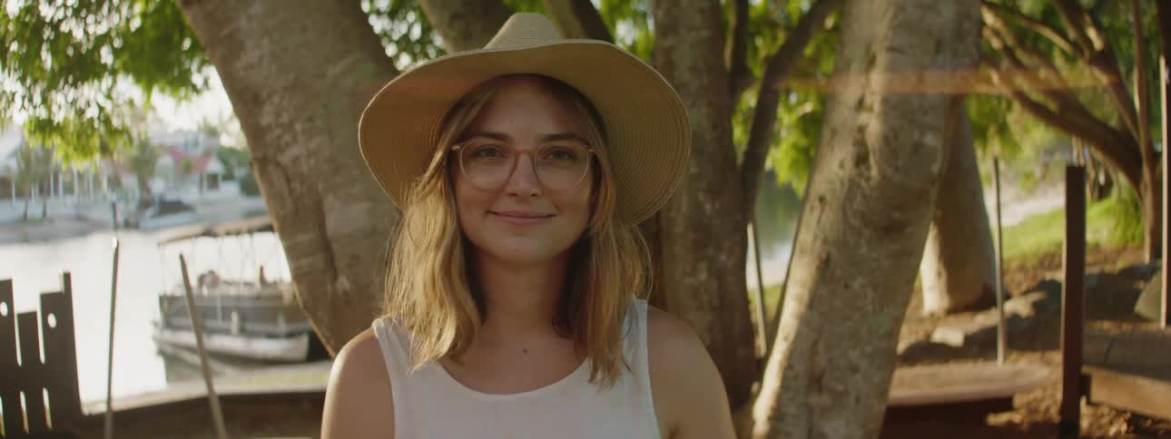 A woman wearing glasses and a sunhat is standing near a tree along the shore as she looks up and smiles