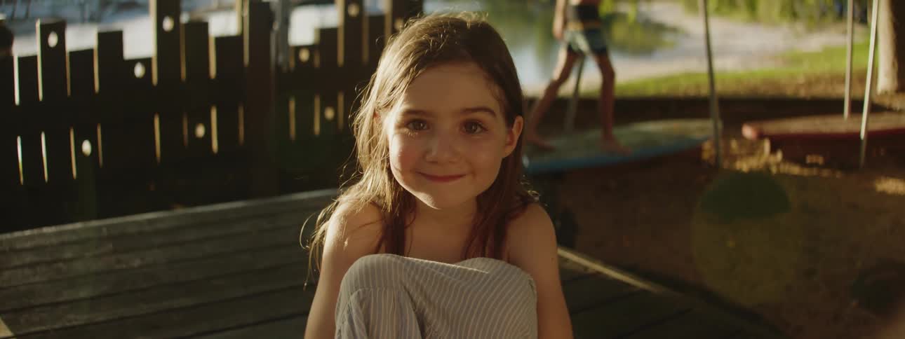 A young girl smile for the camera as she sits outdoors on a wooden bench
