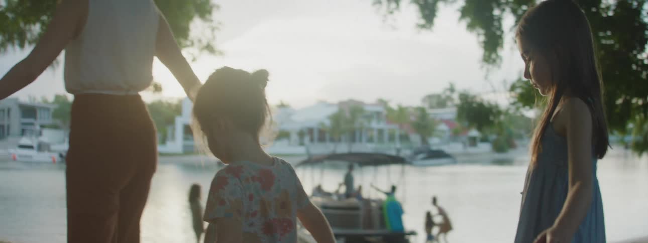 A scene where two little kids are walking towards their mom near a sea where a boat is sailing