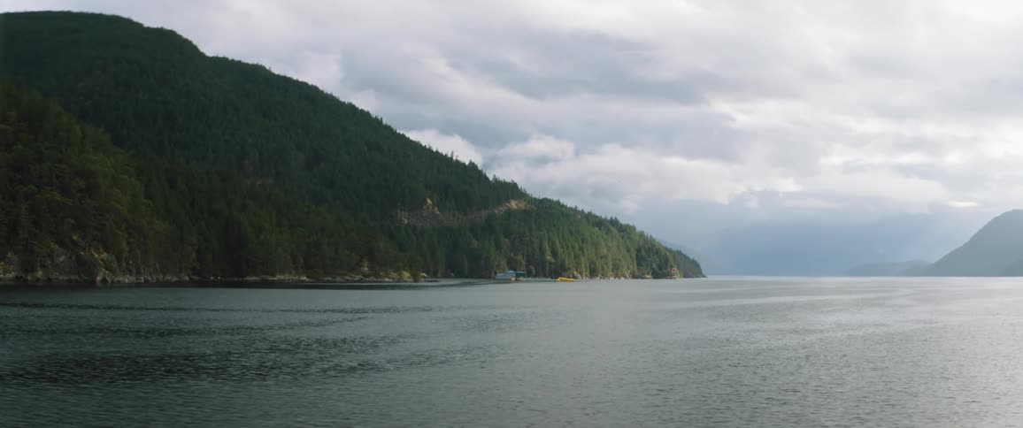 Calm lake waters near the pine forest covered mountains on an overcast day