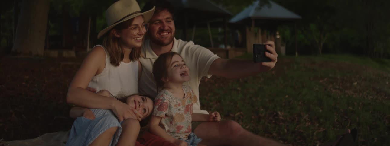 A family of four sits together while posing for a selfie using the dads phone