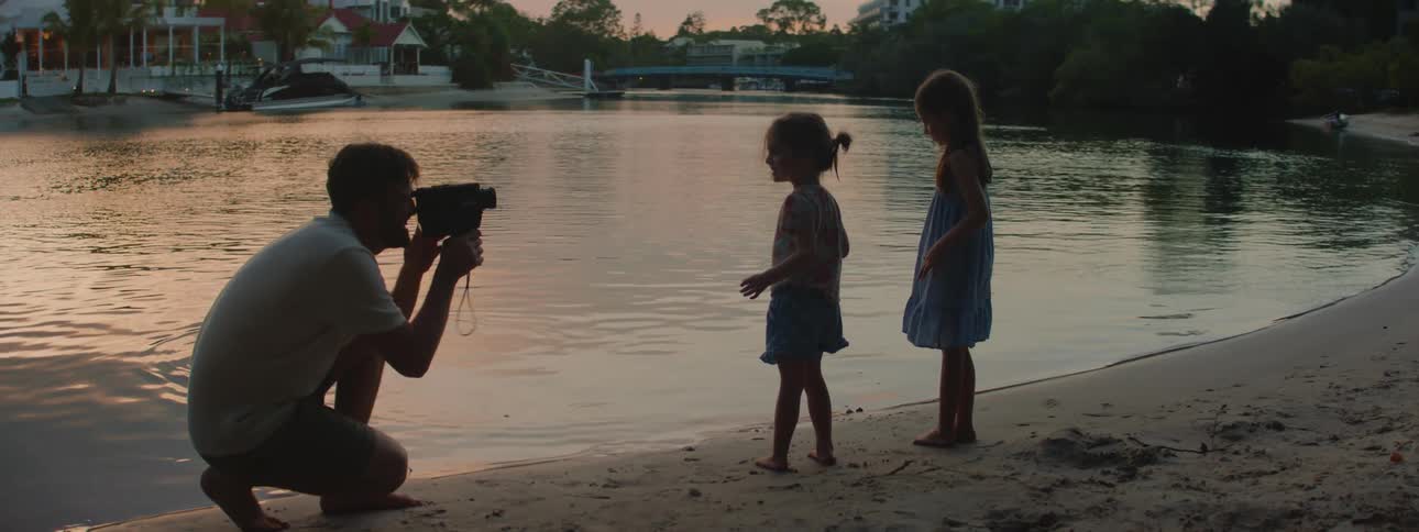 Dad taking home movies of his two young daughters standing on the lake shore little girl is waving at sunset