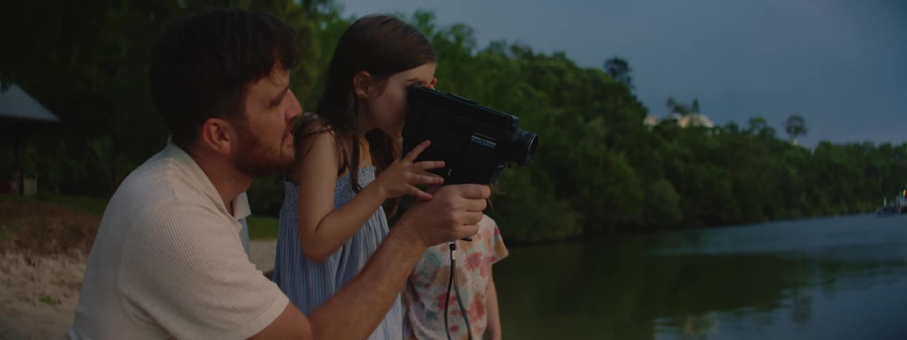 A father teaches his young daughter to use a video camera while his younger son watches