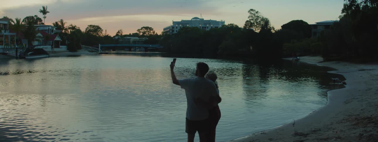 The silhouette of a man and woman standing on the waters edge taking a picture with their cell phone