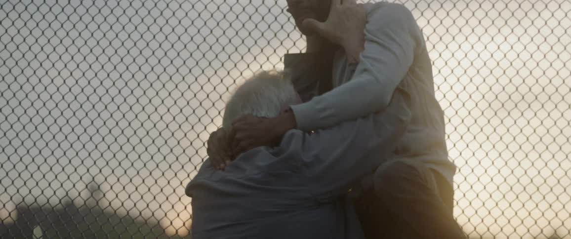 An old man chokes a younger man against a chain link fence