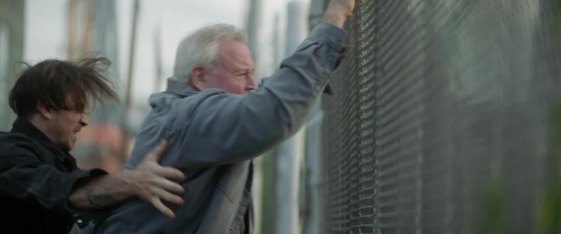 An older man hits a chain link fence with a grimace as another man spins him around
