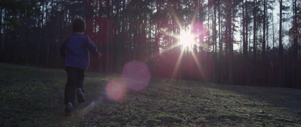 Young boy running towards the sunlight filtering through the tree line at sunset