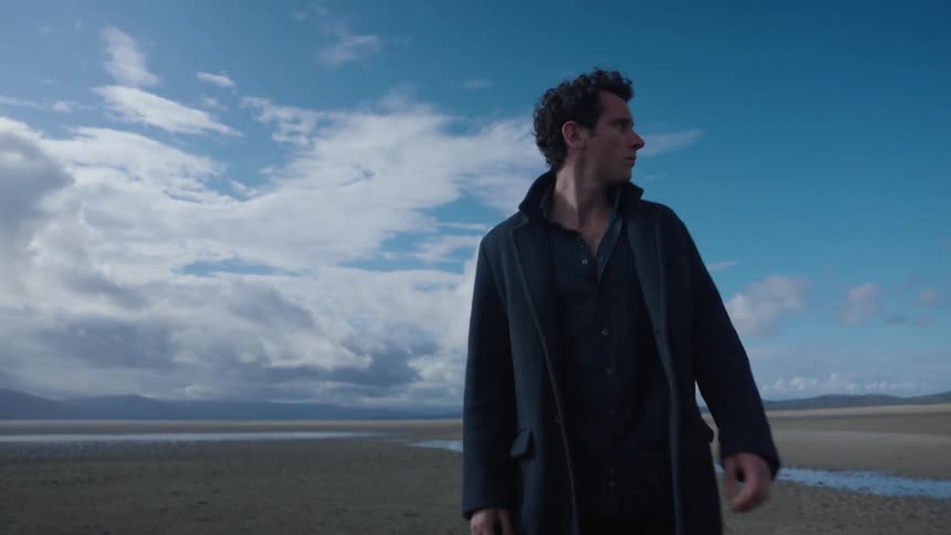 Man wearing coat looks around as he walks across ground near wet sand with mountains in the background