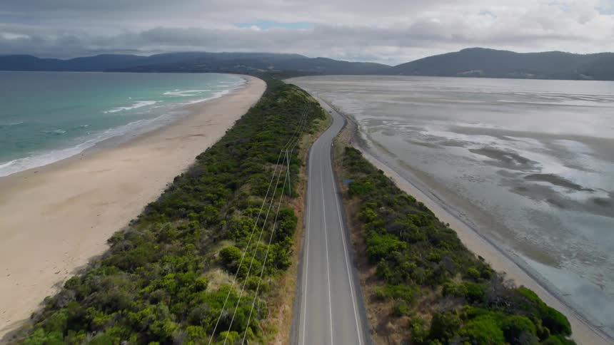 Power lines and highway move along strip of land between beach and mud flats leading toward mountains on cloudy day