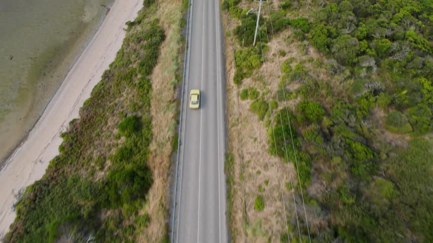 Yellow car parallels power lines while driving past brush and the beach
