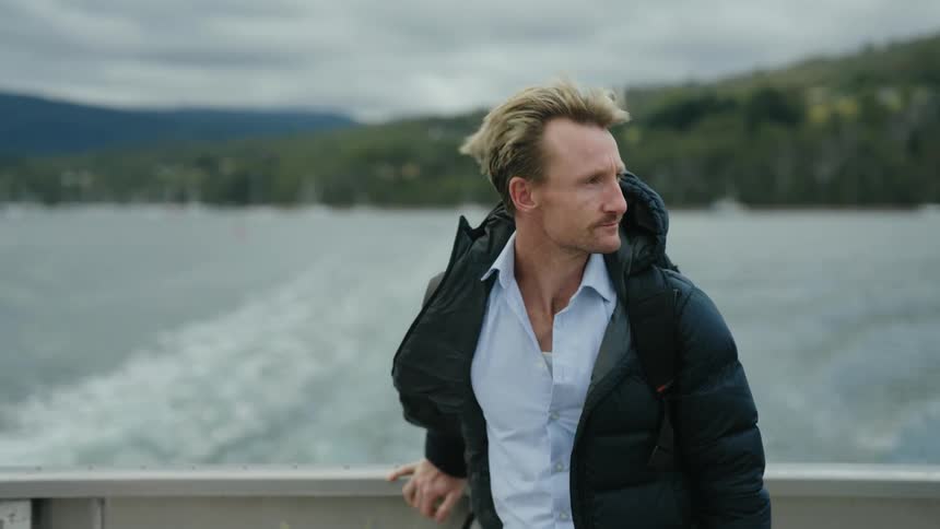 A man in a jacket and backpack stands at the rear end of a boat moving away from a green coast