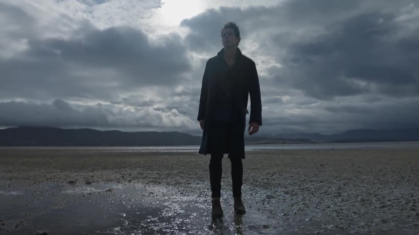 Man walks across damp field toward mountains with brief stop to look behind him