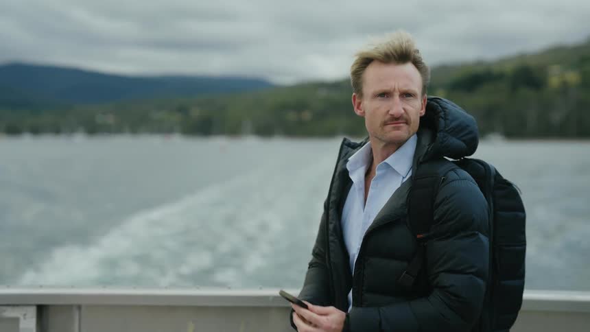 A man wearing a coat and checking his phone while standing on the back of a boat that is navigating on a lake