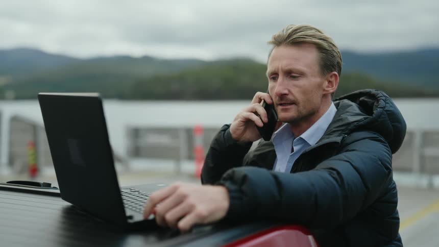 A man talks on the phone while using a laptop propped on his car