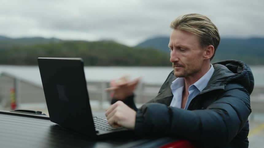 A man standing outdoors along the waterfront opens a laptop and begins typing