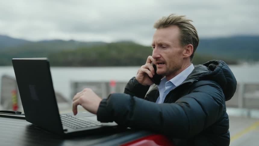 Man standing near truck parked near water makes smartphone and consults laptop