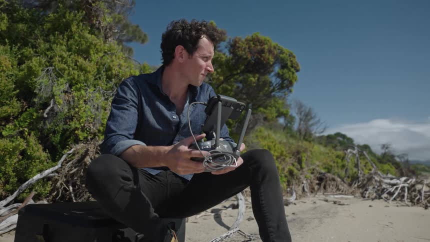Man sits on beach operating drone with mountains on the horizon